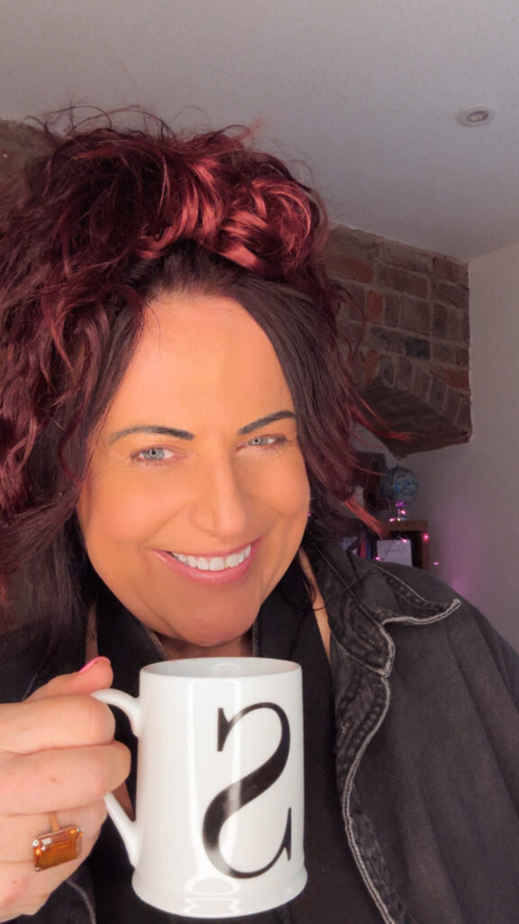 Woman with red curly hair in a cosy jumper standing in morning light holding a mug, representing lazy girl reinvention and calm anti-burnout living.